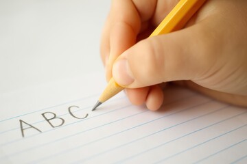 Close-up of a child's hand writing ABC with a pencil on lined notebook paper during handwriting practice