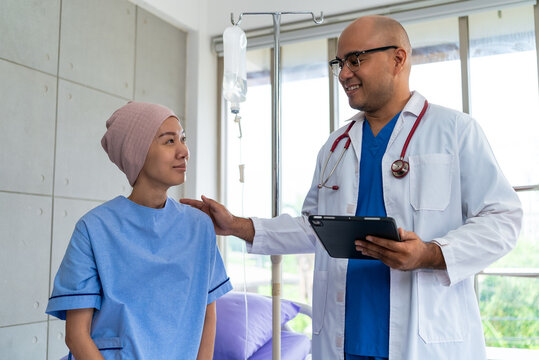Asian woman patient describes her symptoms to an oncologist while sitting on a bed and looking at a man doctor in a lab coat in a hospital ward, health and medical care concept