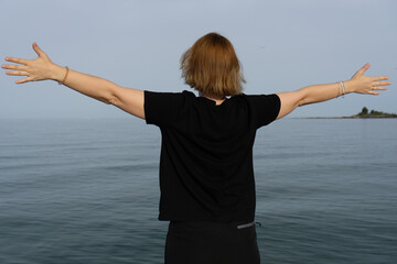 A woman in a black t-shirt and black shorts stands with arms outstretched towards the calm blue sea...