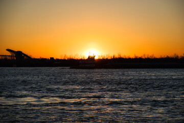 A ship drifts along the Mississippi River, silhouetted against the glowing orange and gold hues of the setting sun. Rippling water catches the light, reflecting New Orleans' quiet farewell to day.