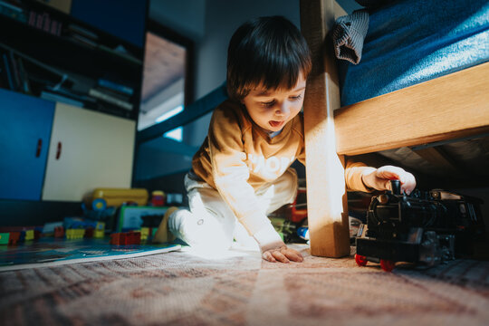 A toddler explores under a bed, excitedly pushing a vintage-style black toy train amidst scattered toys in a brightly lit and warm indoor environment.