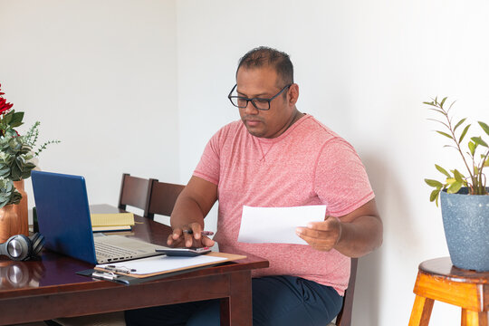 A man in a red shirt is sitting at a table with a laptop and a stack of papers