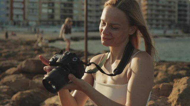 Professional blonde photographer reviewing digital images on rocky coastline, capturing warm seaside landscape with vibrant golden hour colors. Travel, blogging, weekend, relax and lifestyle concept