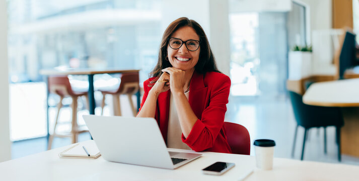 Confident businesswoman in red jacket smiling while working on a laptop - Powered by Adobe