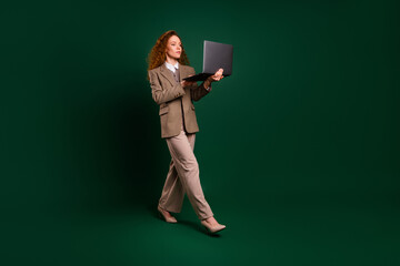 Confident businesswoman walking stylishly with a laptop on an emerald green background in formal...