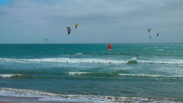 Kitesurfers ride ocean waves near Pacifica, California, along Highway 1. Colorful kites fill the cloudy sky over the turquoise sea in this action-packed coastal scene