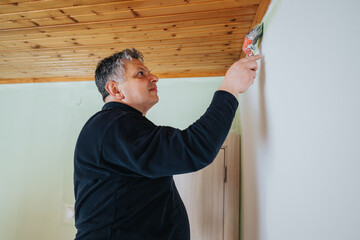 Man diligently paints the edge of a wall near a wooden ceiling using a brush, suggesting home...