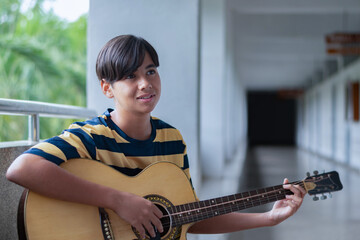 Teen boy playing acoustic guitar, wearing a yellow shirt, looking down to guitar thoughtfully. Creative youth expression and musical lifestyle in a casual setting.