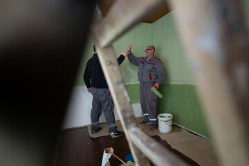 Two workers exchange thumbs-up gestures while painting a room with light green walls, highlighting...