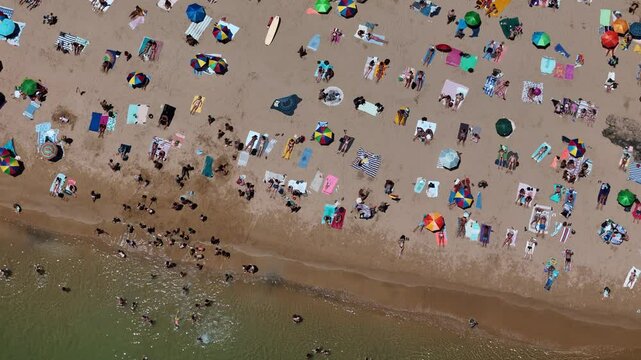 Aerial drone top down view of a crowded beach with colorful umbrellas and sunbathers during a summer afternoon near the coast