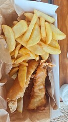 Crispy golden fish and thick-cut fries served in a paper-lined box on a wooden table. Concept of...