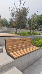 Modern wooden bench stands empty in a tiered concrete urban park surrounded by young trees. Concept of public space design, relaxation, and contemporary city landscaping