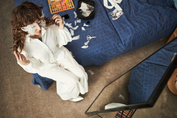 Caucasian young adult woman sitting on bed creating feathered accessory while looking into mirror,...