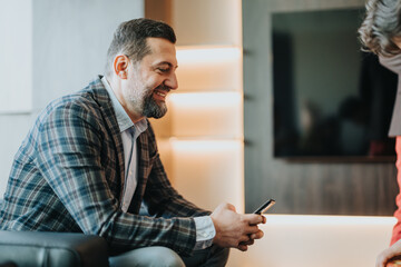 Businessman seated in an office lounge area, holding a smart phone and conversing, illustrating...