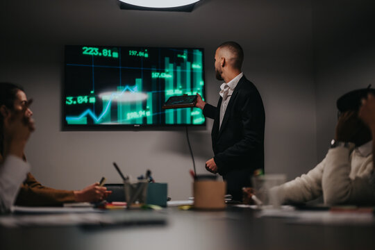 A professional discussing financial trends with colleagues in a corporate meeting room setting. The room features a prominent screen displaying stock market charts, emphasizing financial concepts