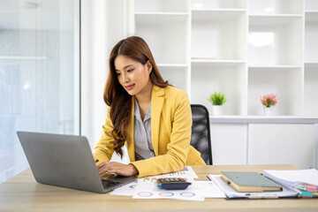 A businesswoman is working in her office. She is checking documents at her desk with a calculator and a mobile phone. She is looking at the calculator and thinking.

