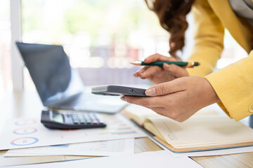 A businesswoman is working in her office. She is checking documents at her desk with a calculator and a mobile phone. She is looking at the calculator and thinking.

