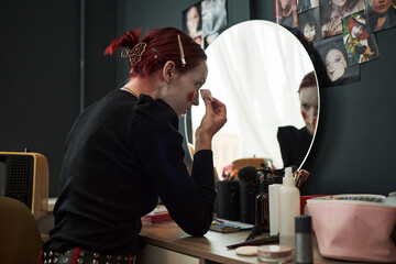 Caucasian teenager girl applying creative makeup in front of mirror, sitting at vanity table with cosmetics, hair clips in hair, photos of makeup looks on wall, focusing on artistic expression