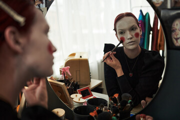 Caucasian teenager girl applying face makeup and red blush with brush in front of mirror, engaging in creative self expression at home, surrounded by cosmetics and personal items