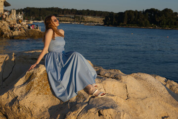 A woman in a blue dress and sunglasses enjoys the golden hour light while sitting on sun-drenched...