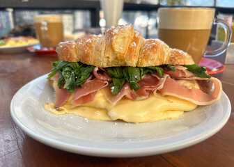 Close-up of a croissant sandwich filled with grilled cheese, ham or bacon, and fresh arugula, served on a wooden table at a street café. Accompanied by a cappuccino or coffee with milk.