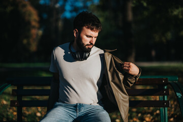 A man relaxes on a park bench, wearing a white shirt and headphones. Sunlight casts shadows as he...