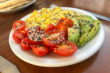 Close-up of a healthy breakfast served in a café or restaurant setting: omelet, sliced tomato, and seed-topped avocado on a plate, on a wooden table.