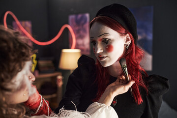 Caucasian teenage girl with red hair sitting indoors having face painted by another teenage girl,...