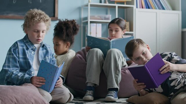 Four multiethnic schoolchildren in casualwear sitting relaxedly on rugged floor in modern classroom setting, studying books during after-class teamwork activity
