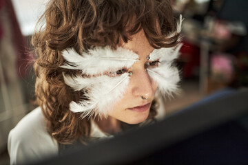 Caucasian teenager girl wearing creative feather makeup sitting indoors, looking at computer...