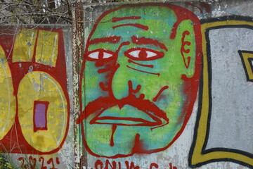 Drawings, inscriptions, pictures, handwriting, signatures. Images made with a can of paint on a brown reinforced concrete wall. A green and yellow face of a man with a mustache drawn from red stripes,