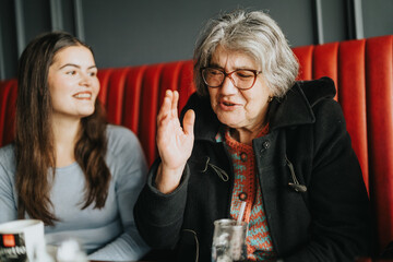 A cheerful elderly woman wearing glasses and a coat shares a lively conversation with a young lady...