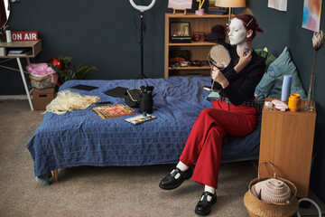 Caucasian teenager girl sitting on bed applying makeup to face using handheld mirror, makeup...