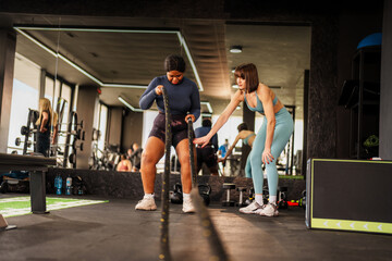 Overweight woman performs a battle ropes exercise with the guidance of a personal trainer,...