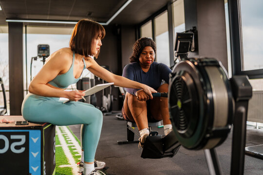 Personal trainer assisting an overweight client during a workout session on a rowing machine, providing guidance and support for achieving fitness goals