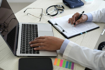 Young professional male doctor is taking notes in journal, physician therapist practitioner filling medical documents with mobile phone or laptop, patient form, illness history, prescription in clinic