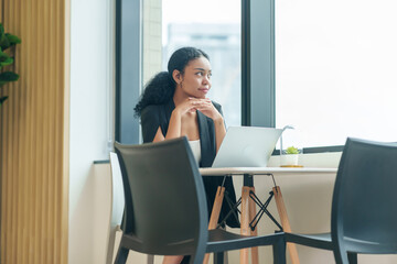 portrait smiling business woman sitting by the window looking away while casual working with laptop in the office room