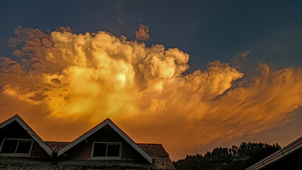 A dramatic golden-orange cloud formation glows during sunset above dark rooftops and distant trees,...