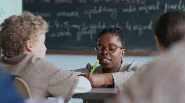 Young Black female teacher with pen in hand guiding young students at desk during classwork, explaining material and being open for discussion, with chalkboard in blurred background
