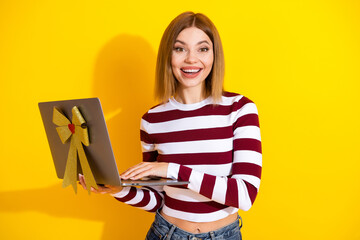 Young woman holding a laptop decorated with a gift bow on a yellow background, expressing...