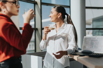 Two business coworkers are taking a coffee break, chatting and smiling in a bright modern office....