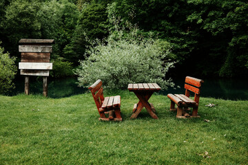 Wooden benches and a table stand on green grass by a calm river, surrounded by dense trees. A...
