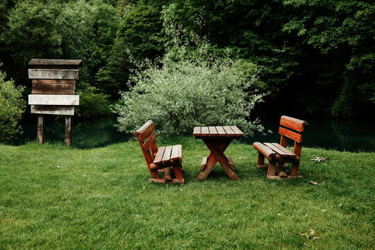 Wooden benches and a table stand on green grass by a calm river, surrounded by dense trees. A rustic signpost adds charm to the secluded spot. Krupajsko vrelo in Serbia.