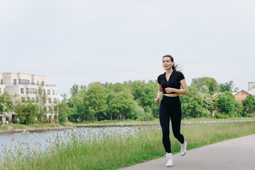 young slim beautiful caucasian woman running on path on river embankment in city park, black...