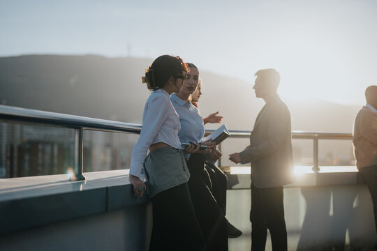 A multicultural group of colleagues engaged in brainstorming on a high tower balcony. The sunset provides a warm backdrop, highlighting collaboration and teamwork among the diverse business people