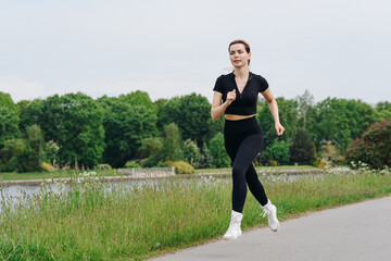 young slim beautiful caucasian woman running on path on river embankment in city park, black...