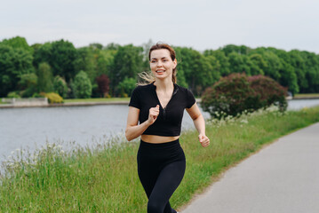 young slim beautiful caucasian woman running on path on river embankment in city park, black...