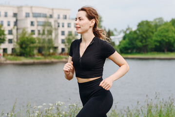 young slim beautiful caucasian woman running on path on river embankment in city park, black...