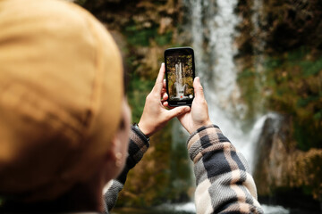 Fototapeta premium Female tourist in plaid jacket filming Veliki Buk waterfall with smartphone at Lisine reserve, Serbia. Travel moment captured in nature. Travel blogger making video outdoor. Close back view.