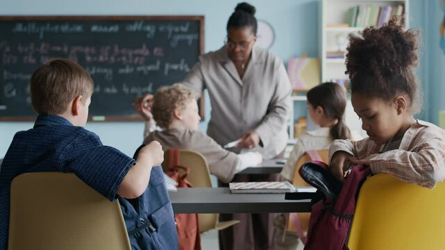 Back view shot of diverse schoolkids sitting at their desks and organizing their belongings in well-lit classroom, as African American female teacher interacting with front row in background